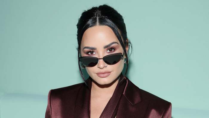 Demi Lovato poses in front of a teal background in a brown satin blazer and sunglasses.